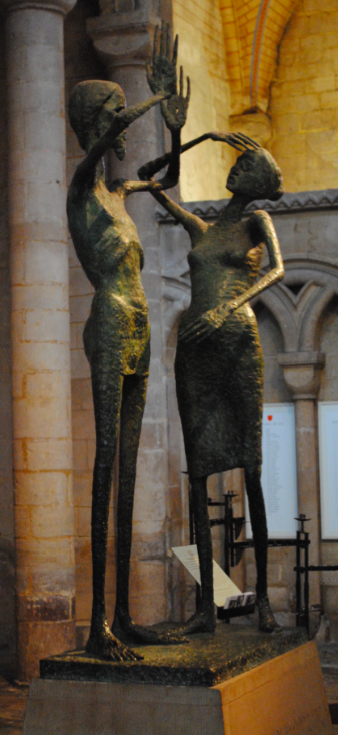 A dark bronze sculpture of two tall, elongated, stylized nude figures — one raising a hand to touch the other's face — stands on a plinth inside a stone cathedral interior with columns and arches.