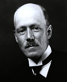 Black-and-white photographic portrait of a balding, mustached man in formal dark dress with a white collar and bow tie.