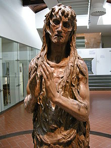 A carved wooden statue depicts a gaunt, hollow-eyed woman with long matted hair draped over her body and hands folded at her chest, displayed in a modern interior with glass walls and tiled floor.