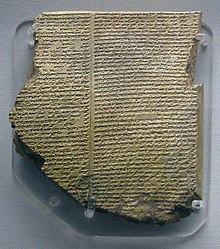Photograph of an ancient inscribed clay tablet covered in cuneiform script, chipped and weathered, displayed upright on a stand.
