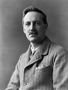 Black-and-white photographic portrait of a mustached man in a tweed jacket with a handkerchief in the breast pocket.