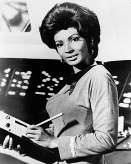 A black-and-white publicity photo of Lt. Uhura (actor Nichelle Nichols) in her Starfleet uniform, writing at a communications console.