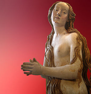 A nude wooden statue of a woman, her body covered chiefly by her own long flowing hair, stands with hands clasped in front of her, photographed against a red backdrop.