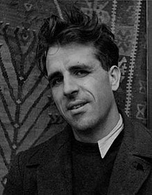 Black-and-white photographic portrait of a dark-haired young man in a dark top with a white collar, seated in front of a patterned wall hanging.