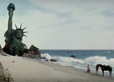 Color film still of a half-buried, weathered Statue of Liberty (torch and crown visible) lying on a beach, with two small human figures and a horse standing on the sand nearby.