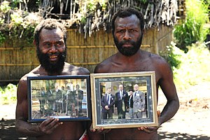 Photograph of two bare-chested men on Tanna island holding up framed photographs of Prince Philip in military dress posing with local tribesmen.