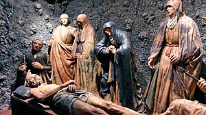 A row of life-size polychrome terracotta figures in hooded robes stand grouped around a nude male figure laid on the ground, photographed in dim gallery lighting.