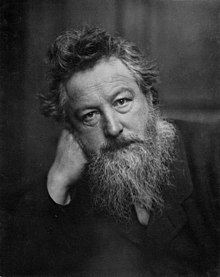 Black-and-white photographic portrait of a bearded, tousle-haired man in a dark coat, resting his head on his hand.